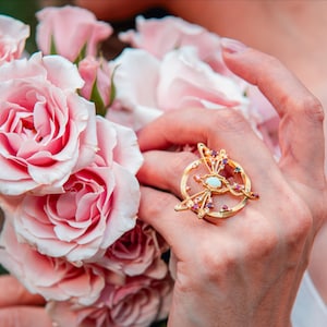May include: A gold ring featuring a circular frame with a central oval opal cabochon, accented by small red rubies and clear white stones arranged in a celestial pattern. The ring has a polished finish and sits against a bouquet of light pink roses.