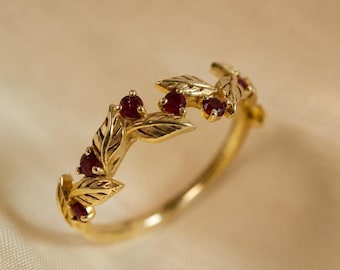 May include: A gold ring featuring a band designed with sculpted leaves and small, round, deep red rubies set between the foliage. The metal has a polished finish, and the botanical motif wraps partially around the circumference of the ring.