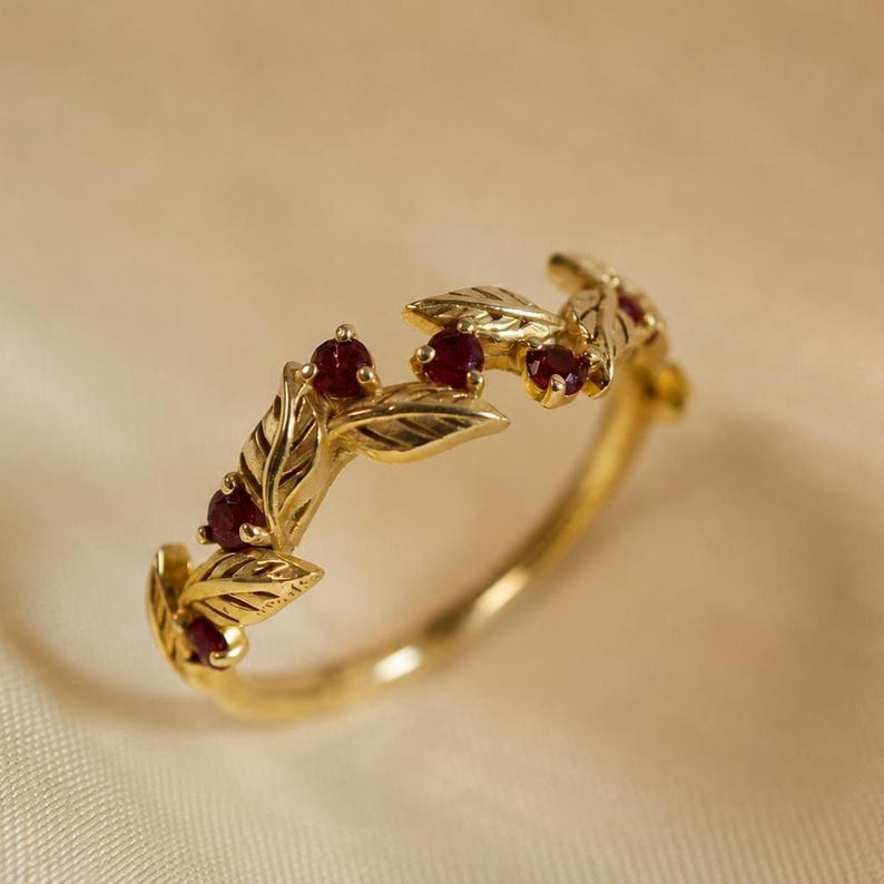 May include: A gold ring featuring a band designed with sculpted leaves and small, round, deep red rubies set between the foliage. The metal has a polished finish, and the botanical motif wraps partially around the circumference of the ring.