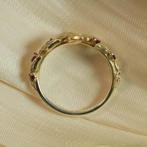 May include: A gold ring featuring a series of small, round, deep red gemstones set into a band with a decorative, organic, leaf-like metal pattern. The band is circular and polished, showcasing a warm metallic tone and multiple red accents.