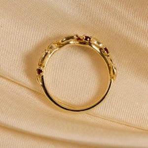 May include: A gold ring featuring a series of small, round, deep red gemstones set along the upper portion of the band. The metal band has a smooth, polished finish and a circular shape, showcasing a delicate design with multiple stone settings.