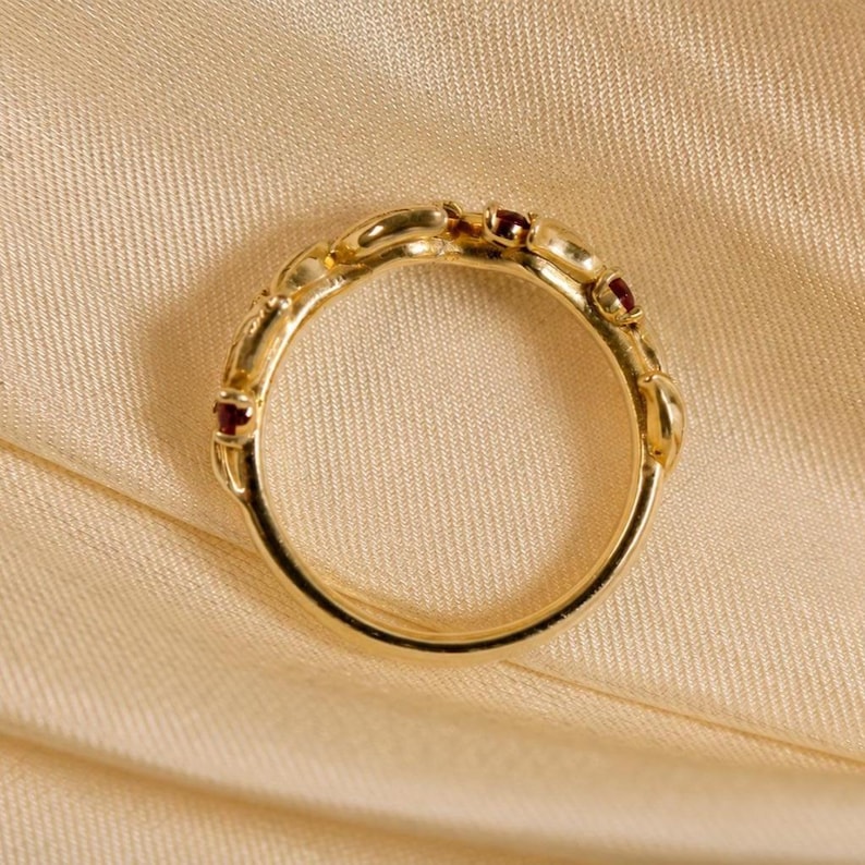 May include: A gold ring featuring a series of small, round, deep red gemstones set along the upper portion of the band. The metal band has a smooth, polished finish and a circular shape, showcasing a delicate design with multiple stone settings.