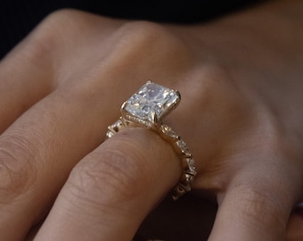 Peut inclure: Une bague en or avec une grosse pierre précieuse claire de forme rectangulaire au centre. La monture est également ornée de petites pierres précieuses claires. La bague est portée au doigt.