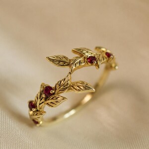 Puede incluir: Un anillo de oro con un delicado motivo de enredadera con hojas esculpidas y pequeños rubíes redondos de color rojo intenso engastados en el metal. La banda está hecha de oro amarillo pulido, mostrando texturas de hojas intrincadas y un diseño botánico.