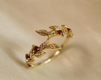 May include: A gold ring featuring a delicate vine motif with sculpted leaves and small, round, deep red rubies set within the metal. The band is crafted from polished yellow gold, showcasing intricate leaf textures and a continuous botanical design.