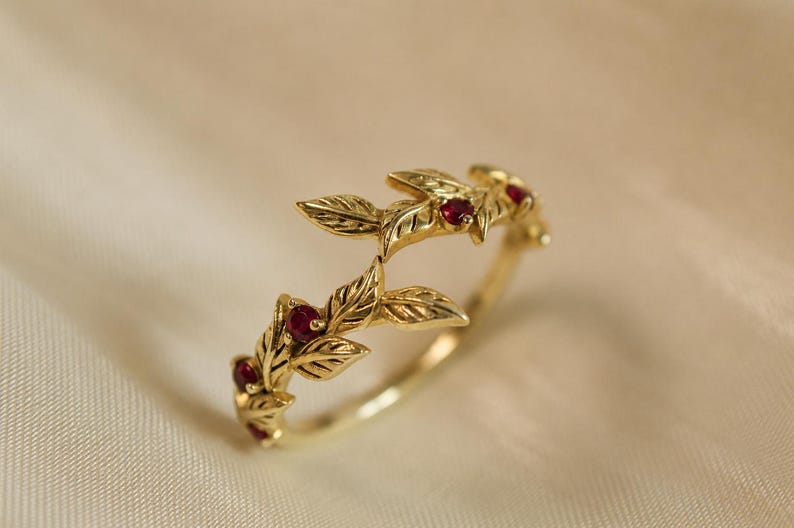 May include: A gold ring featuring a delicate vine motif with sculpted leaves and small, round, deep red rubies set within the metal. The band is crafted from polished yellow gold, showcasing intricate leaf textures and a continuous botanical design.