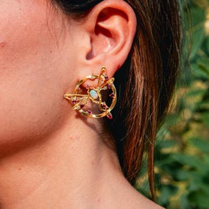 May include: A gold-toned circular earring featuring a central iridescent opal stone surrounded by a delicate wire-frame butterfly motif. The design is accented with small, vibrant red gemstones embedded along the circular frame and butterfly wings.