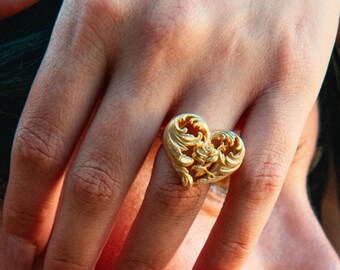 May include: A gold-toned ring featuring an intricate heart-shaped design with openwork filigree detailing and a central rose motif. The metal surface has a matte finish, and the heart shape is oriented with the point facing downward on the finger.