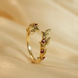 May include: A gold open-style ring featuring a delicate vine motif with sculpted leaves and four small, round, deep red gemstones set along the band. The ring has a polished finish and an adjustable gap between the two ends of the vine design.