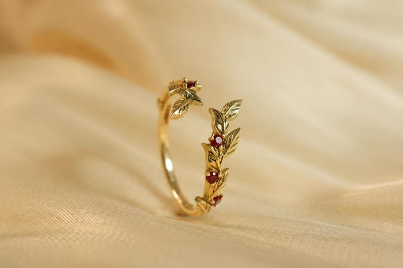 May include: A gold open-style ring featuring a delicate vine motif with sculpted leaves and four small, round, deep red gemstones set along the band. The ring has a polished finish and an adjustable gap between the two ends of the vine design.