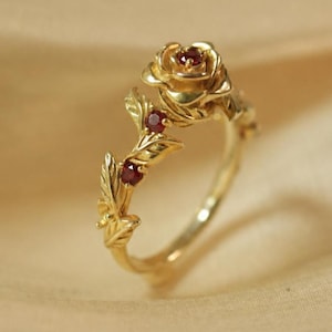 May include: A gold ring featuring a sculpted rose at the top and a vine-like band with leaves. The center of the rose and two leaves are accented with small, round, deep red rubies. The metal has a polished finish and a warm yellow gold tone.
