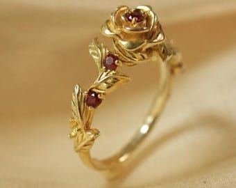 May include: A gold ring featuring a sculpted rose at the top and a vine-like band with leaves. The center of the rose and two leaves are accented with small, round, deep red rubies. The metal has a polished finish and a warm yellow gold tone.
