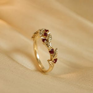 May include: A gold ring featuring a band designed with sculpted leaves and small, round, deep red gemstones set between the foliage. The metal has a polished finish, and the design wraps around the upper portion of the circular band.