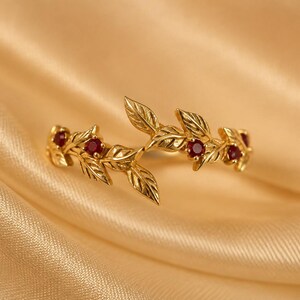 May include: A gold ring featuring a delicate branch design with sculpted leaves and four small, round, deep red rubies set within the metal foliage. The band is open at the center, showcasing a detailed botanical motif with polished gold surfaces.