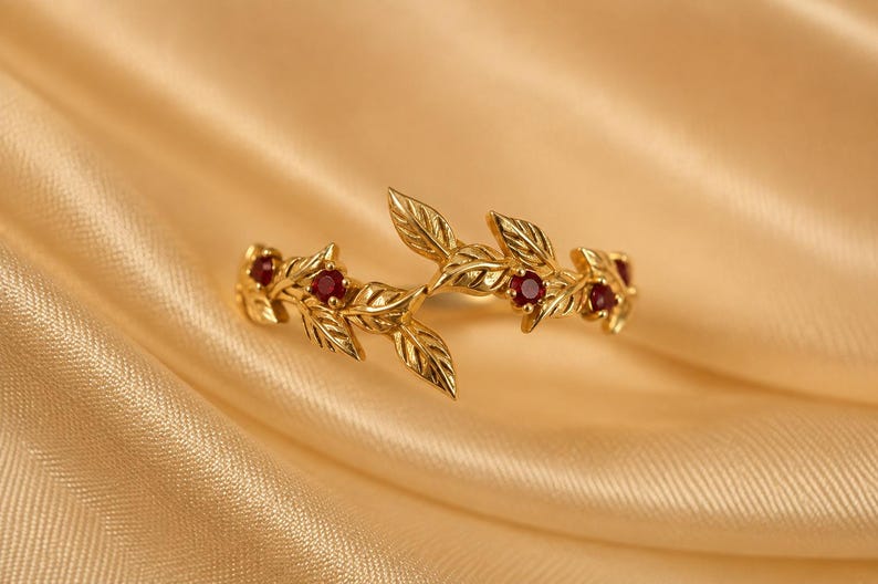May include: A gold ring featuring a delicate branch design with sculpted leaves and four small, round, deep red rubies set within the metal foliage. The band is open at the center, showcasing a detailed botanical motif with polished gold surfaces.