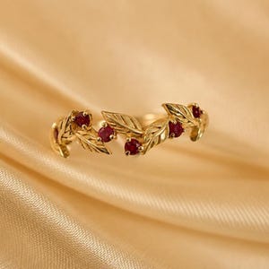 May include: A gold ring featuring a band designed with interconnected metal leaves and four small, round, deep red rubies set between the foliage. The metal has a polished finish, and the design wraps around the finger in a continuous pattern.