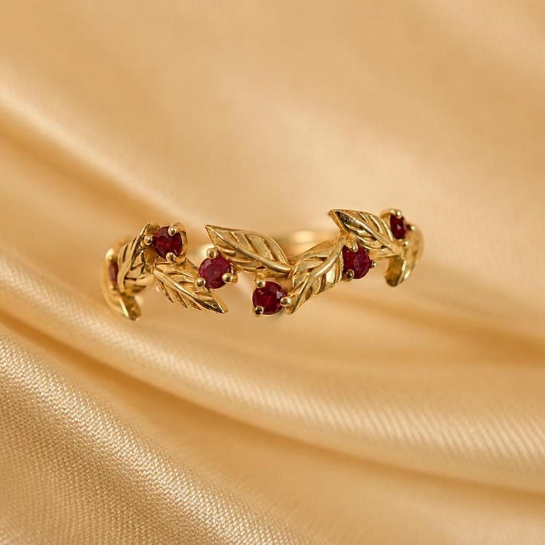 May include: A gold ring featuring a band designed with interconnected metal leaves and four small, round, deep red rubies set between the foliage. The metal has a polished finish, and the design wraps around the finger in a continuous pattern.