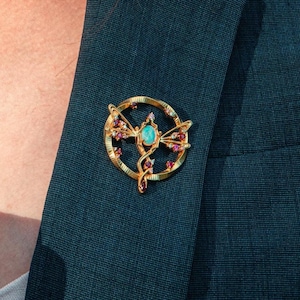 May include: A gold-toned circular brooch featuring a central oval-shaped light blue stone. The design includes a stylized, elongated figure accented with small, round, deep red stones embedded along the frame and the central vertical element.