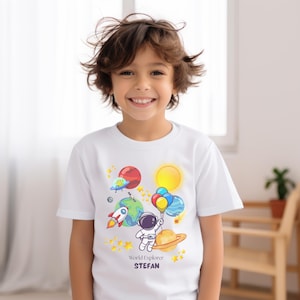 Op de afbeelding: Wit T-shirt met een kleurrijk ruimtethema. Het ontwerp bevat planeten, een raket, een astronaut met ballonnen en de tekst "World Explorer STEFAN". Het shirt wordt gedragen door een lachend kind.