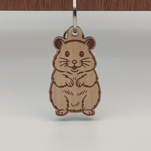May include: A wooden keychain featuring a smiling hamster design. The keychain is light brown with etched details, including whiskers, paws, and fur. It has a silver-colored metal keyring attached at the top, perfect for keys or bags.