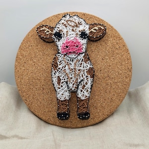 May include: String art of a cow on a corkboard. The cow is made with brown, white, and black thread, with a pink nose and black eyes. The artwork is mounted on a round corkboard, offering a rustic and charming appearance.