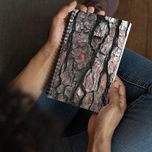 May include: A spiral-bound notebook featuring a detailed, close-up image of tree bark. The bark displays a textured pattern with shades of brown, gray, and reddish-brown. The notebook is held open, showcasing the bark design prominently.