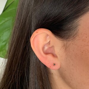 K&ouml;nnte beinhalten: Nahaufnahme eines Ohrs mit einem kleinen schwarzen Ohrstecker. Der Ohrstecker ist in einer goldenen Fassung gefasst.