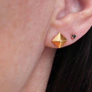 K&ouml;nnte beinhalten: Ein Paar goldfarbene Ohrstecker. Die Ohrringe sind geometrisch geformt und &auml;hneln einer Pyramide. Ein Ohrring hat einen kleinen schwarzen Edelstein in der Mitte.