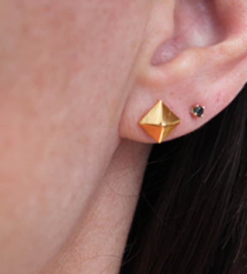 K&ouml;nnte beinhalten: Ein Paar goldfarbene Ohrstecker. Die Ohrringe sind geometrisch geformt und &auml;hneln einer Pyramide. Ein Ohrring hat einen kleinen schwarzen Edelstein in der Mitte.