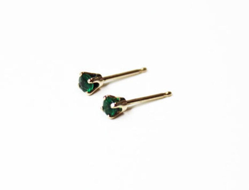 May include: A pair of gold stud earrings with small, round, emerald green gemstones.