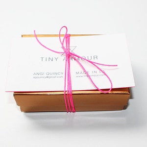 May include: A small, gold box tied with a pink ribbon. The box has a white label with the text "Tiny Armour" and contact information for Angie Quincy.