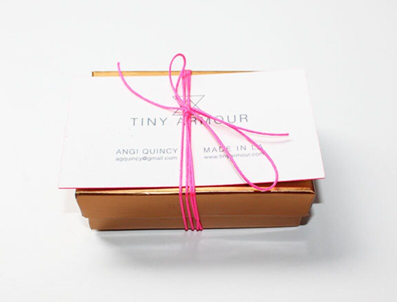 May include: A small, gold box tied with a pink ribbon. The box has a white label with the text "Tiny Armour" and contact information for Angie Quincy.