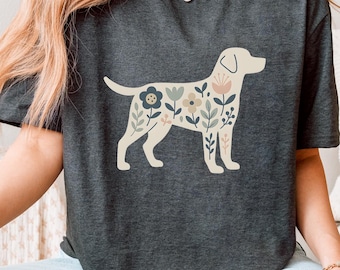 May include: Dark gray t-shirt featuring a cream-colored dog silhouette filled with floral designs in muted tones of blue, pink, and green. The shirt has a crew neck and short sleeves. The overall theme is a nature-inspired design.