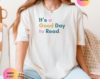 May include: A white t-shirt with the text "It's a Good Day to Read." in colorful letters. The shirt is being worn by a person sitting on a couch.