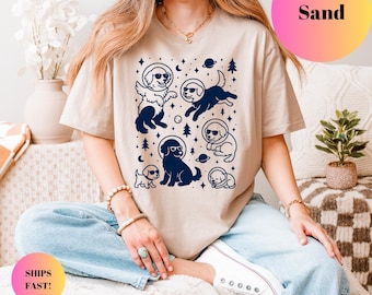 May include: A sand-coloured t-shirt with a navy blue graphic of dogs in space suits, planets, and stars. The shirt has short sleeves. The word "Sand" is printed on the top right corner of the image.