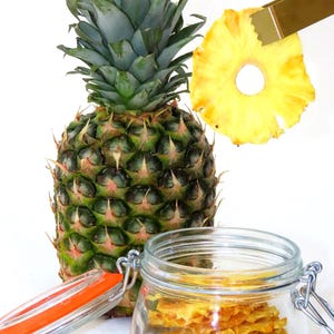 May include: A whole pineapple with green leaves and a jar filled with dried pineapple slices. The dried pineapple slices are light yellow and translucent.