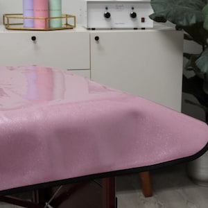 May include: A pink glitter spa table cover with a black border. The cover is made of a waterproof material and is designed to protect the table from spills and stains.