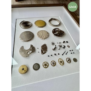 Puede incluir: Una exhibici&oacute;n enmarcada de piezas de relojes antiguos, incluyendo engranajes, muelles y esferas. La disposici&oacute;n presenta una variedad de componentes met&aacute;licos, con un fondo blanco y un marco de madera. El texto "12x16" es visible.