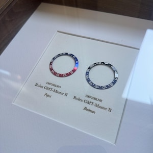 May include: A framed display featuring two Rolex GMT-Master II watch bezels. One is red and blue, the other black and blue. The red and blue bezel is labelled "Pepsi", the black and blue bezel is labelled "Batman."