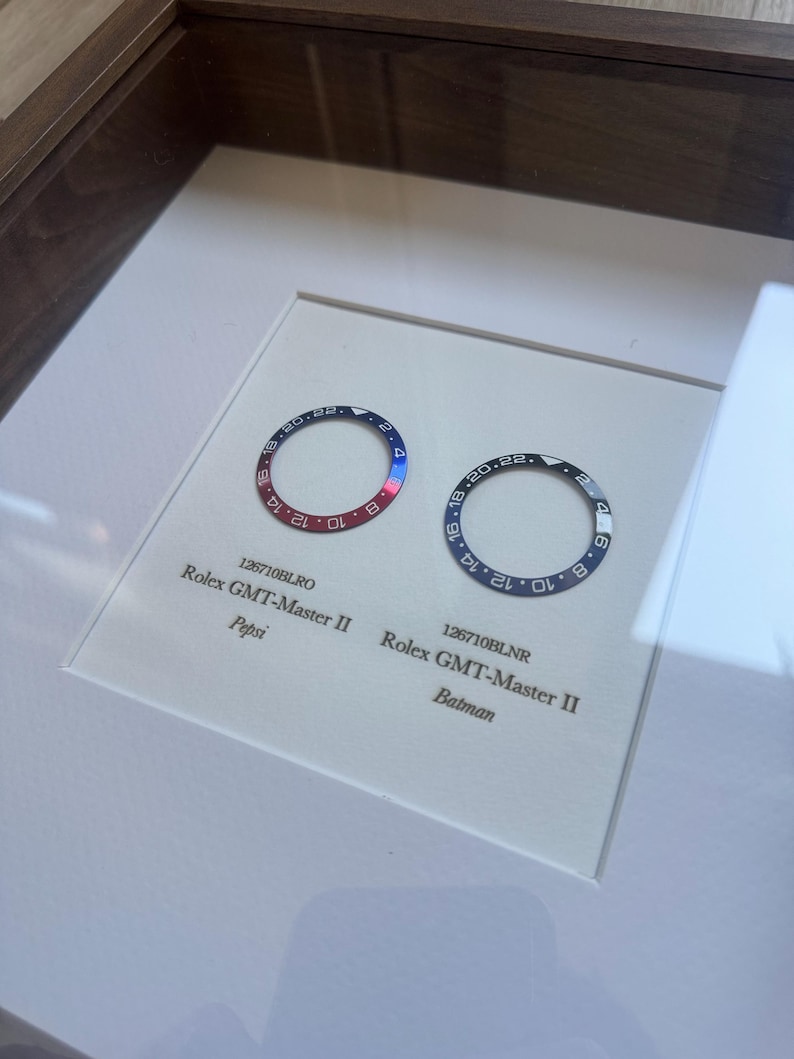 May include: A framed display featuring two Rolex GMT-Master II watch bezels. One is red and blue, the other black and blue. The red and blue bezel is labelled "Pepsi", the black and blue bezel is labelled "Batman."
