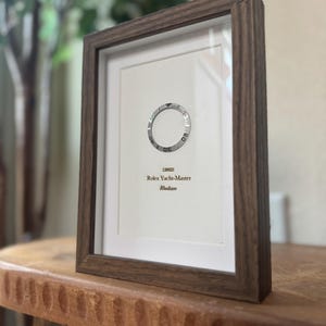 May include: Framed print featuring a Rolex Yacht-Master watch bezel. The print is in a dark wood frame and includes the text "126622 Rolex Yacht-Master Rhodium". The frame is displayed on a wooden surface.