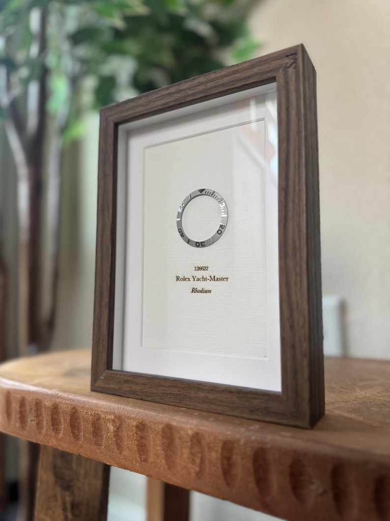 May include: Framed print featuring a Rolex Yacht-Master watch bezel. The print is in a dark wood frame and includes the text "126622 Rolex Yacht-Master Rhodium". The frame is displayed on a wooden surface.