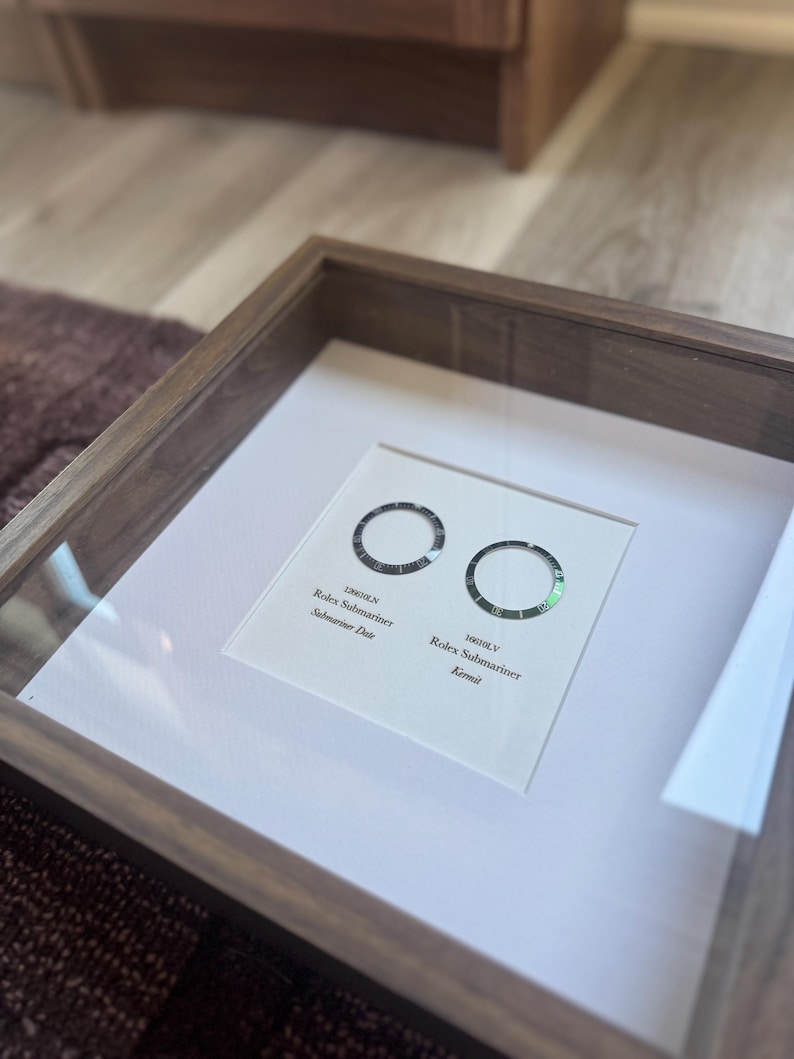May include: A wooden framed display showcasing two Rolex Submariner watch bezels. The left bezel is labelled "126610LN Submariner Date", the right one "16610LV Kermit". The frame is made of dark wood.