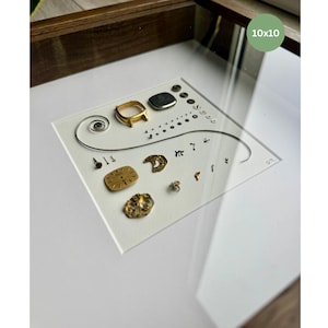 Puede incluir: Una obra de arte enmarcada que presenta componentes de reloj sobre un fondo blanco. La disposici&oacute;n incluye piezas de reloj doradas y plateadas, tornillos peque&ntilde;os y una espiral. El marco es de madera marr&oacute;n oscuro y la obra est&aacute; etiquetada como "10x10" (25,4 x 25,4 cm).