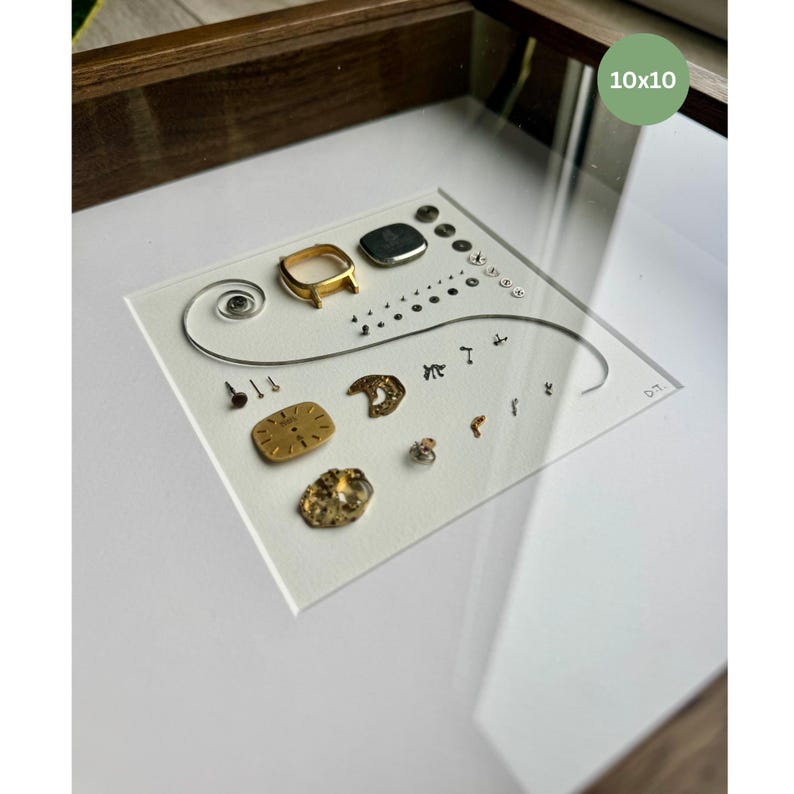 Puede incluir: Una obra de arte enmarcada que presenta componentes de reloj sobre un fondo blanco. La disposici&oacute;n incluye piezas de reloj doradas y plateadas, tornillos peque&ntilde;os y una espiral. El marco es de madera marr&oacute;n oscuro y la obra est&aacute; etiquetada como "10x10" (25,4 x 25,4 cm).