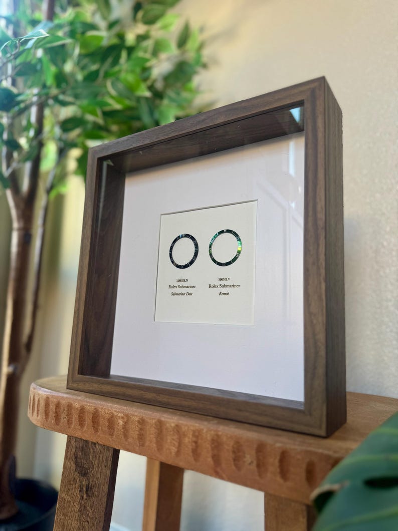 May include: A framed print with two circular designs with iridescent detailing. The dark wood frame surrounds a white mount with text below the circles. The text reads "Rolex Submariner" and "Submariner Date" and "Rome". The frame is on a wooden stool.