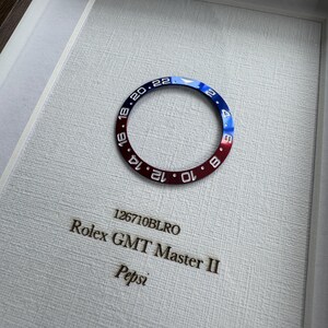 May include: A framed Rolex GMT Master II bezel, featuring a red and blue color scheme with white numerals. The text "126710BLRO" and "Pepsi" are visible. The bezel is set against a textured white background.