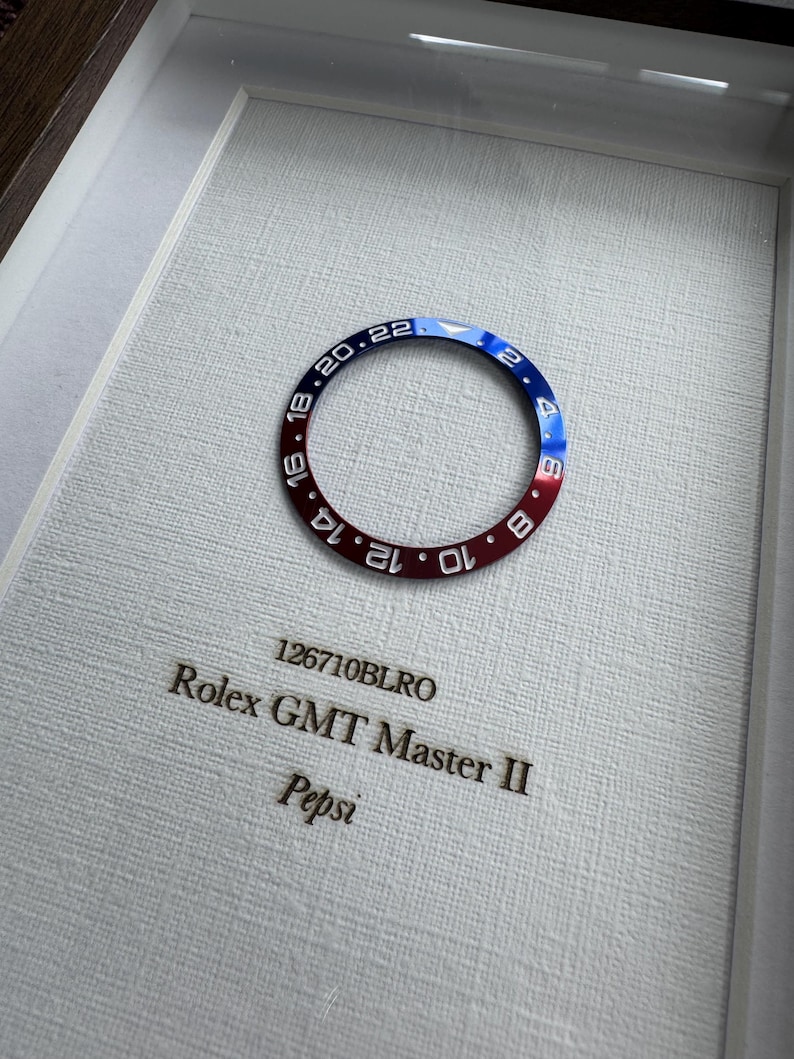 May include: A framed Rolex GMT Master II bezel, featuring a red and blue color scheme with white numerals. The text "126710BLRO" and "Pepsi" are visible. The bezel is set against a textured white background.