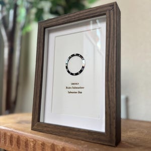 May include: Framed print featuring a watch bezel design. The print includes the text "126610LN Rolex Submariner Submariner Date" in a dark wooden frame. The frame is displayed on a wooden surface.