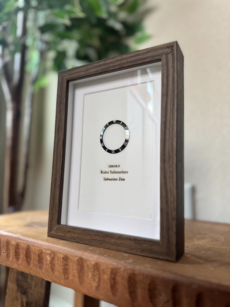 May include: Framed print featuring a watch bezel design. The print includes the text "126610LN Rolex Submariner Submariner Date" in a dark wooden frame. The frame is displayed on a wooden surface.